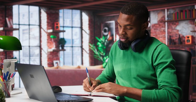 African american male teleworker writing personal notes at his desk in modern loft, remote job. Productive guy sitting at home office, using pen to take notes in his textbook. Camera B.